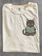 folded ivory Comfort Colors shirt featuring a black cat reading a book chest design, cozy literary shirt for readers, librarians, and cat lovers