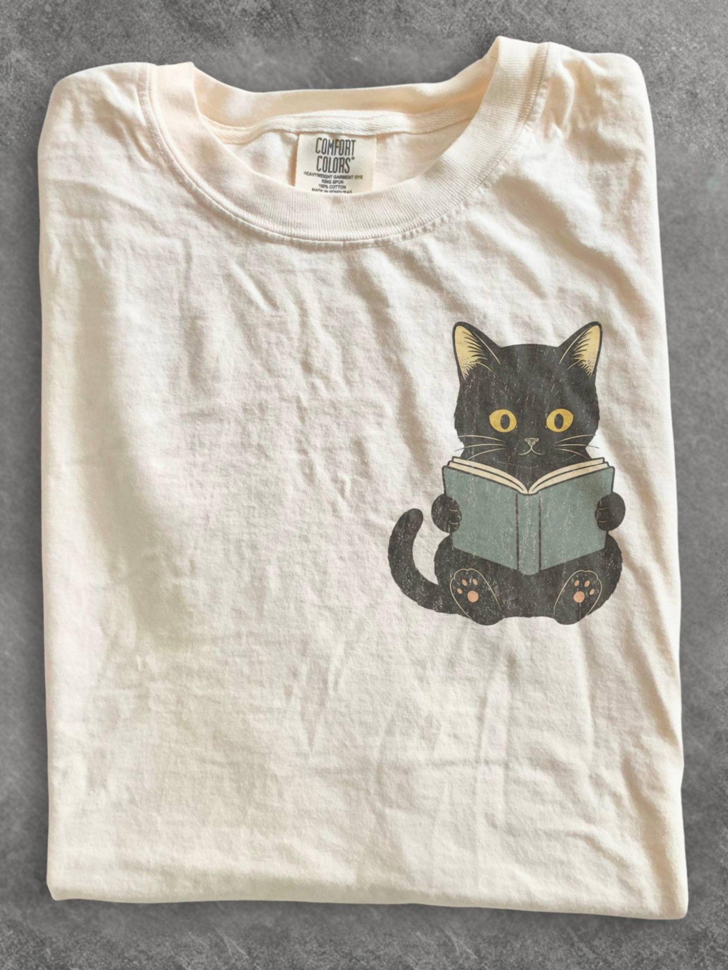 folded ivory Comfort Colors shirt featuring a black cat reading a book chest design, cozy literary shirt for readers, librarians, and cat lovers