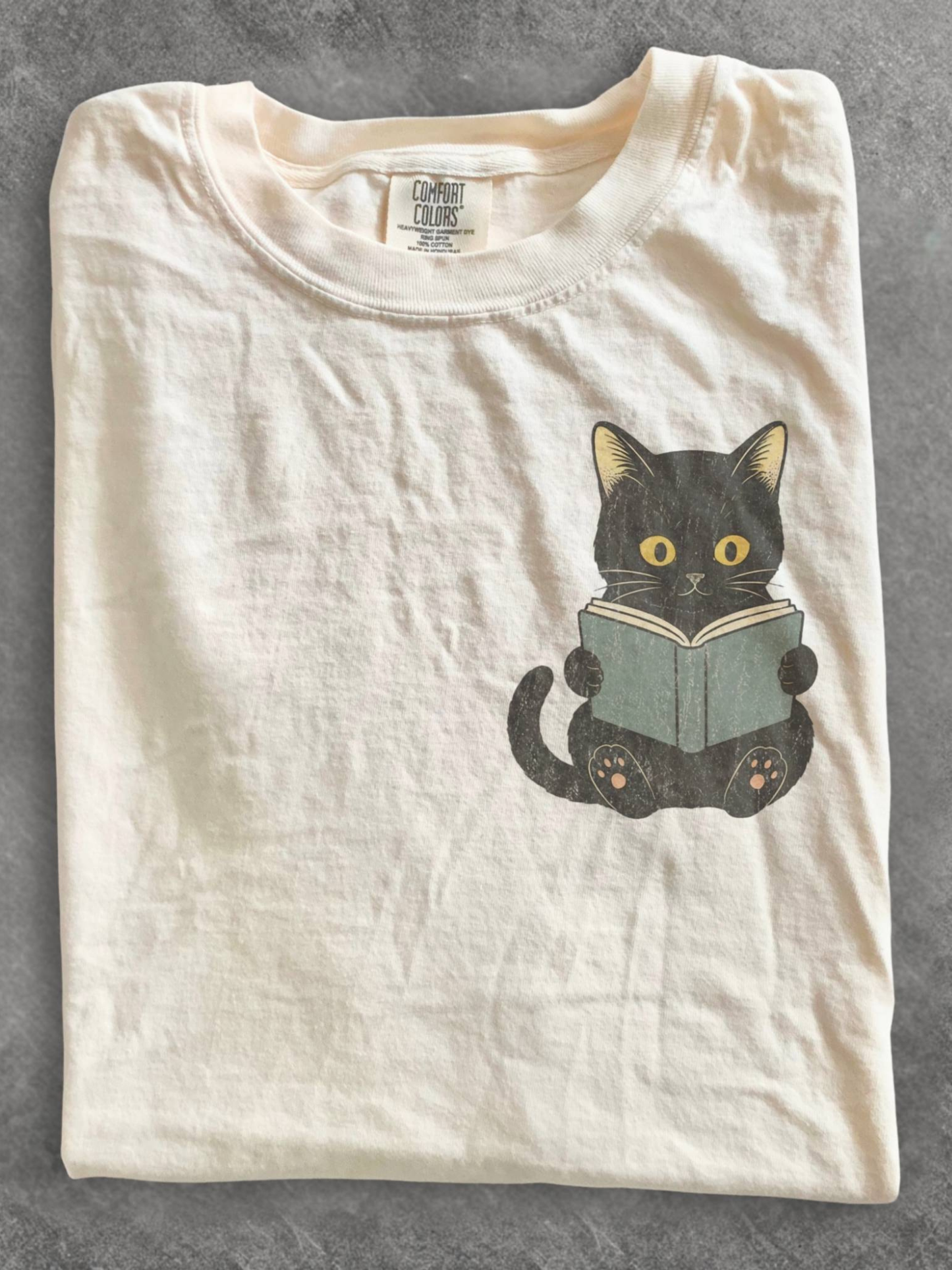 folded ivory Comfort Colors shirt featuring a black cat reading a book chest design, cozy literary shirt for readers, librarians, and cat lovers
