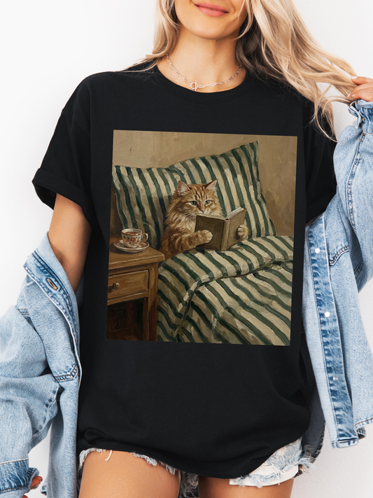Black graphic t-shirt featuring a vintage style illustration of a cat reading a book in bed next to a tea cup, styled with a light denim jacket and layered jewelry. Casual outfit for book lovers and cat lovers who enjoy cozy reading themed apparel.