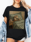 Black graphic t-shirt featuring a vintage style illustration of a cat reading a book in bed next to a tea cup, styled with a light denim jacket and layered jewelry. Casual outfit for book lovers and cat lovers who enjoy cozy reading themed apparel.