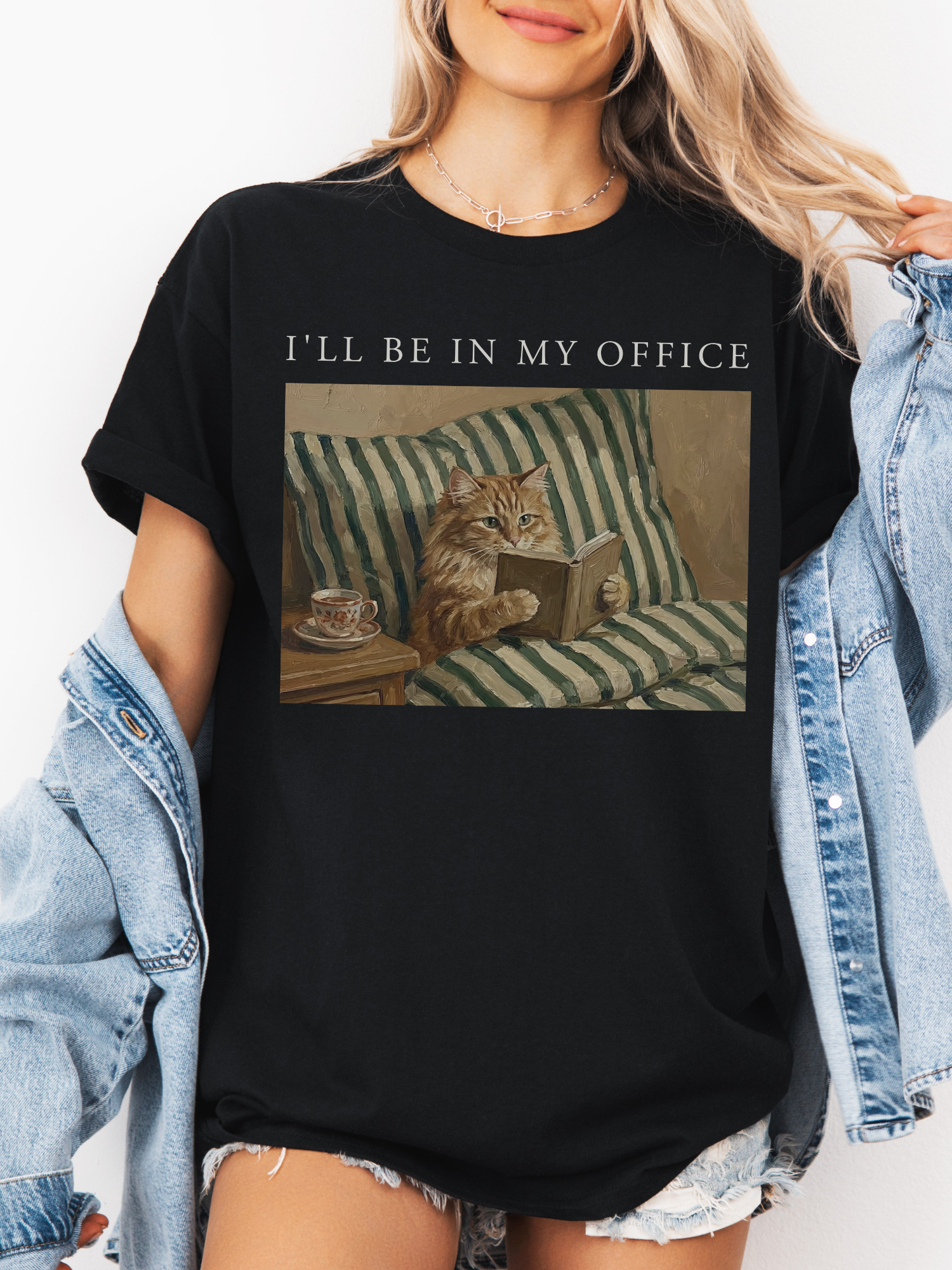 Black oversized t shirt featuring the phrase Ill be in my office above a vintage style illustration of a cat reading a book in bed with a tea cup, styled with a denim jacket. Cozy and funny book lover shirt perfect for readers and cat lovers.