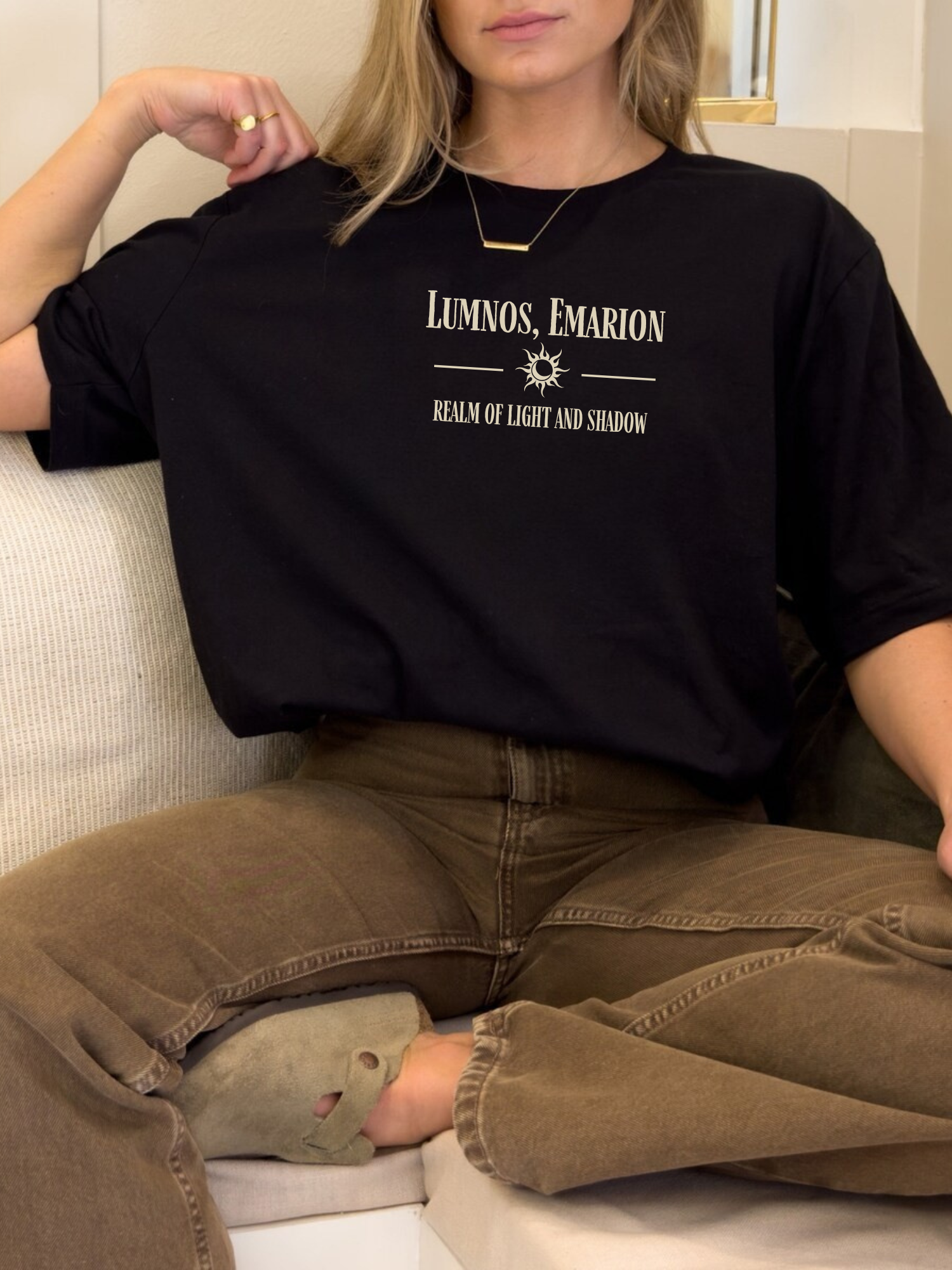 Woman lounging on a couch in a black t-shirt with a fantasy quote design that says "Lumnos, Emarion - Realm of Light and Shadow" in light tan, paired with brown jeans and cozy slippers

