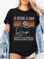 Woman wearing black t shirt with opossum reading book graphic and a book a day keeps the mental breakdown away text styled with denim jacket and casual outfit