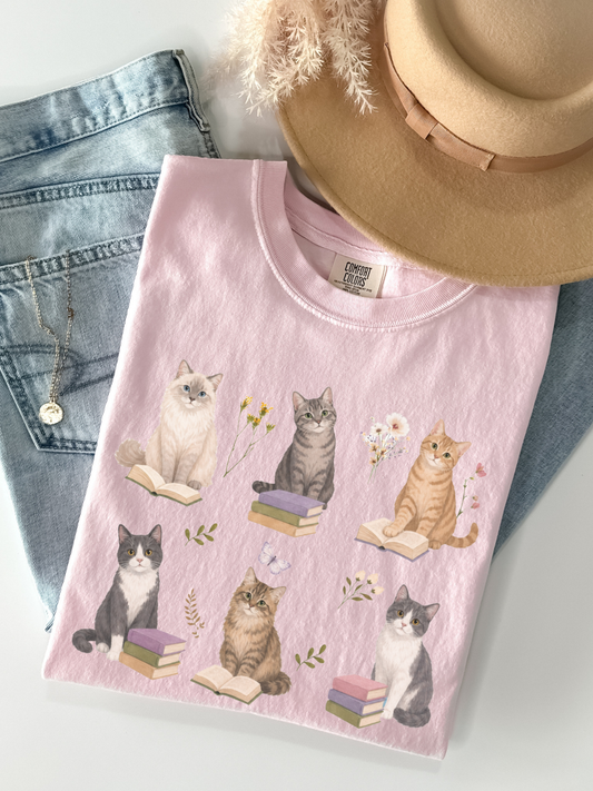 Flat lay of Blossom Comfort Colors t shirt with cute cats sitting on books surrounded by flowers styled with denim jeans necklace and beige hat book lover aesthetic