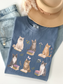 Flat lay of blue jean Comfort Colors t shirt with illustrated cats reading books and floral elements styled with denim jeans gold necklace and beige hat