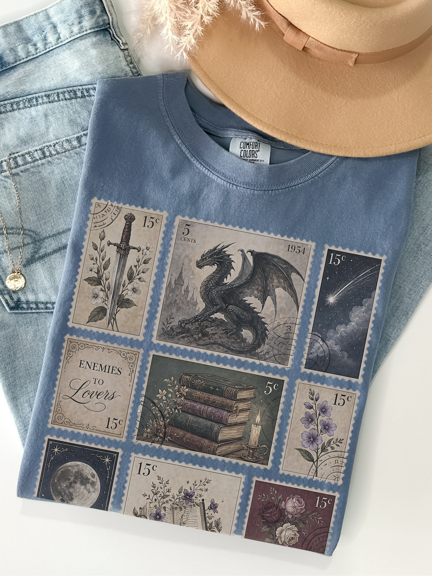 Flat lay of blue jean Comfort Colors fantasy book lover t shirt with vintage postage stamp collage featuring dragon sword moon books flowers castle and enemies to lovers artwork with jeans and hat