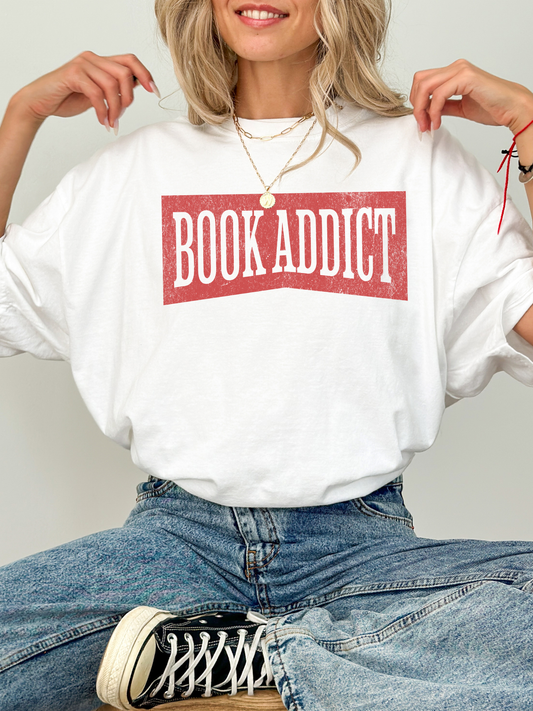 woman sitting cross legged wearing an ivory comfort colors t shirt with distressed red book addict lettering and blue jeans