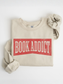 folded sand colored sweatshirt with red vintage book addict text laid flat on a white background