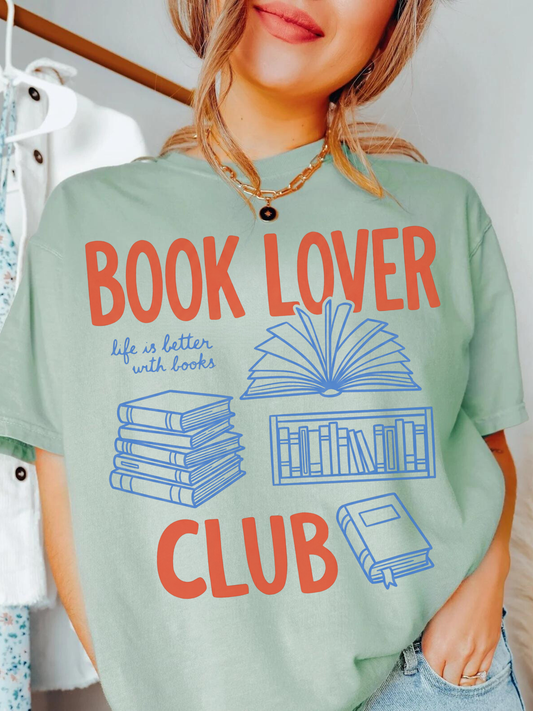 woman wearing a sage green comfort colors t shirt with book lover club text and illustrated books styled casually indoors