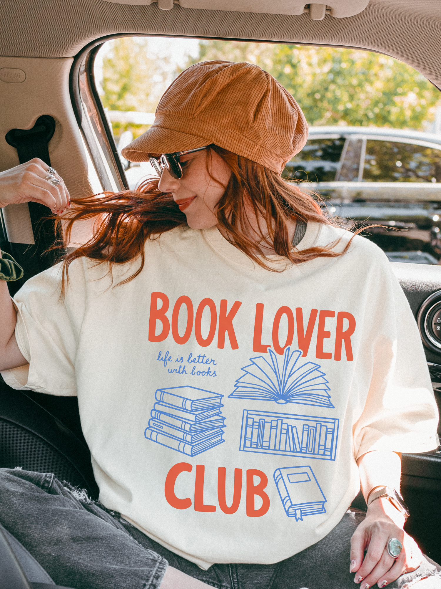 woman sitting in a car wearing an ivory comfort colors t shirt with book lover club design styled with hat and sunglasses