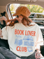 woman sitting in a car wearing an ivory comfort colors t shirt with book lover club design styled with hat and sunglasses