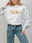 A woman wearing an ash gray crewneck sweatshirt with a colorful autumn tree graphic on the chest, paired with blue jeans, against a plain light gray background.