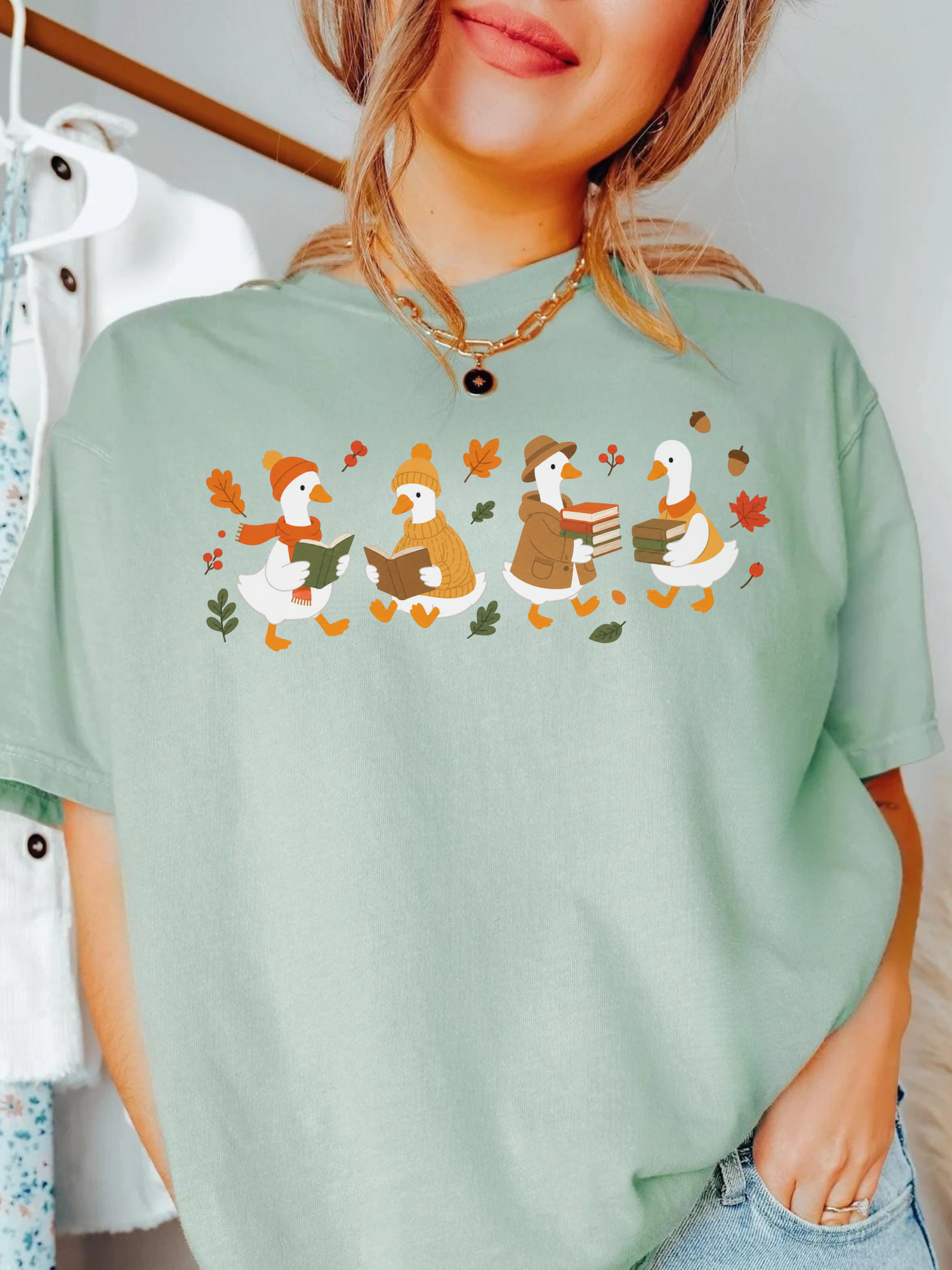 A woman wearing a bay green t-shirt with a graphic of four cartoon ducks in autumn clothing walking with leaves. The background shows a clothing rack with other garments.