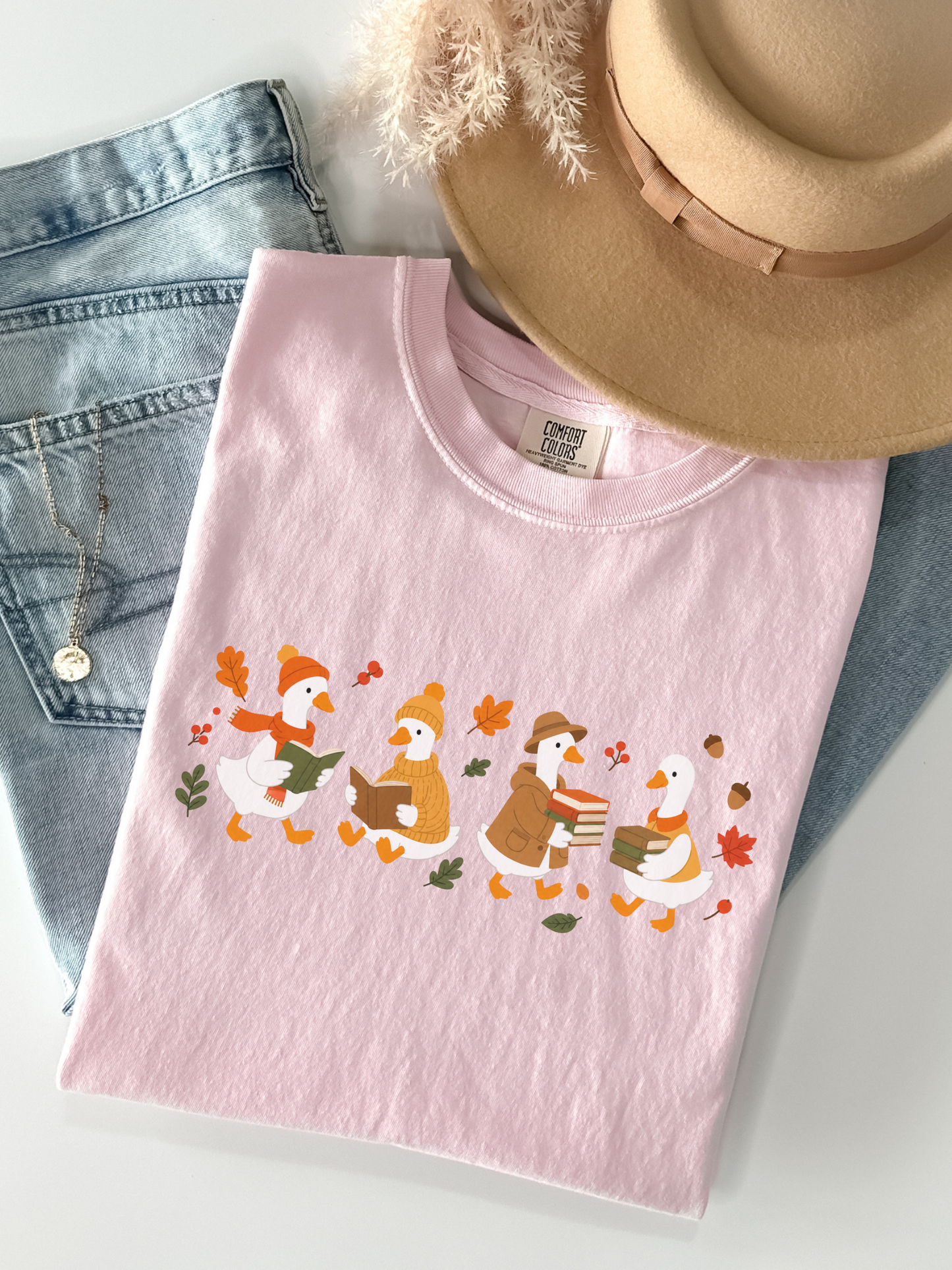 A blossom crewneck t-shirt featuring a graphic of four cartoon ducks dressed in autumn clothing, styled in a flat lay with blue jeans, a tan hat, and a gold necklace on a white surface.