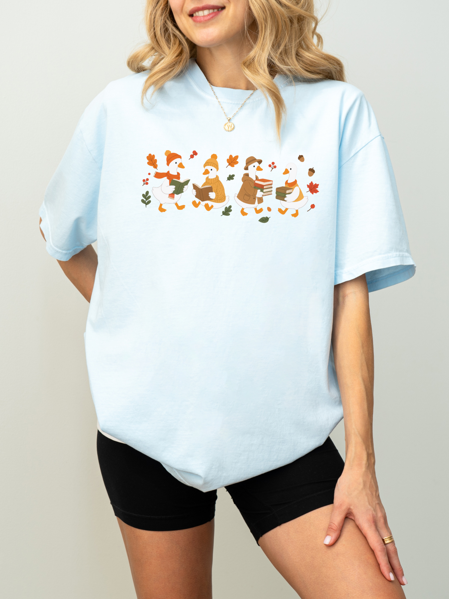 A woman wearing an oversized chambray t-shirt with a colorful graphic of three penguins in winter clothing surrounded by falling leaves, paired with black biker shorts.
