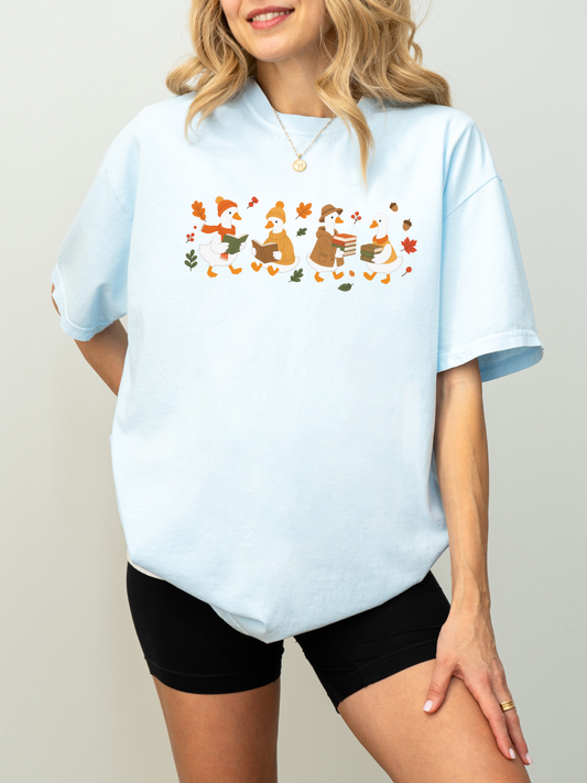 A woman wearing an oversized chambray t-shirt with a colorful graphic of three penguins in winter clothing surrounded by falling leaves, paired with black biker shorts.