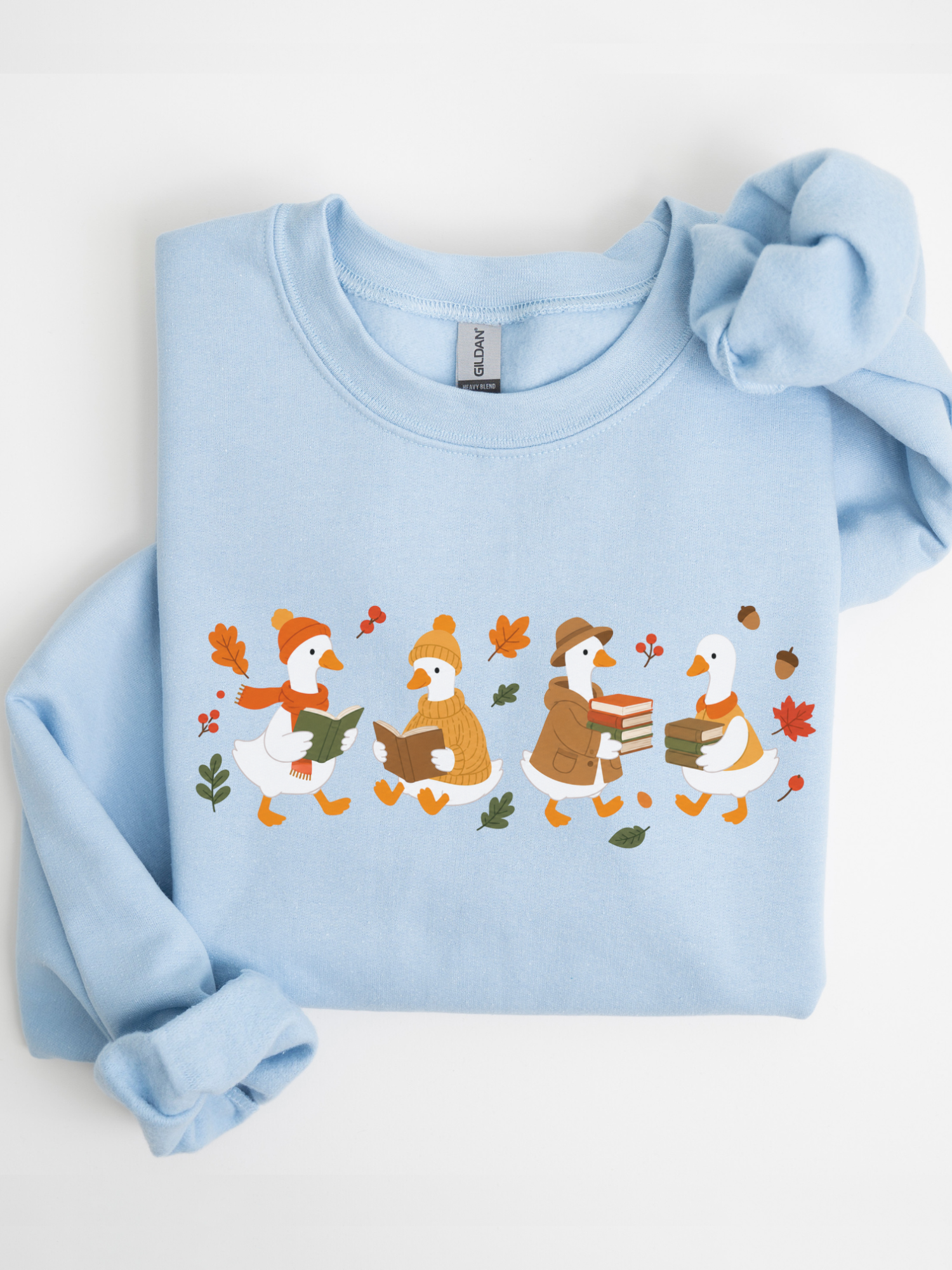 A light blue crewneck sweatshirt featuring a graphic of four cartoon ducks reading books under autumn leaves, styled flat on a white background.