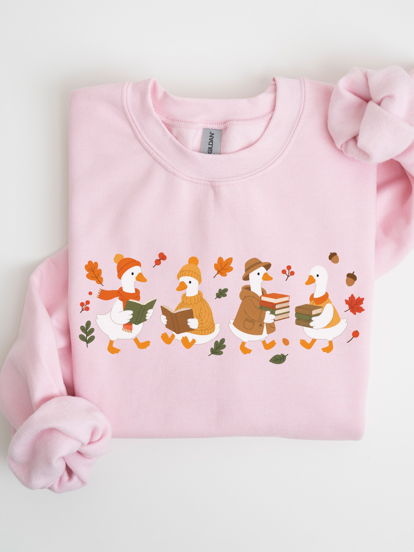 A light pink crewneck sweatshirt featuring a graphic of four cartoon ducks reading books in autumn outfits, surrounded by colorful leaves and acorns. The sweatshirt is neatly folded on a white background with a matching pink fabric bow tied to the upper right corner.