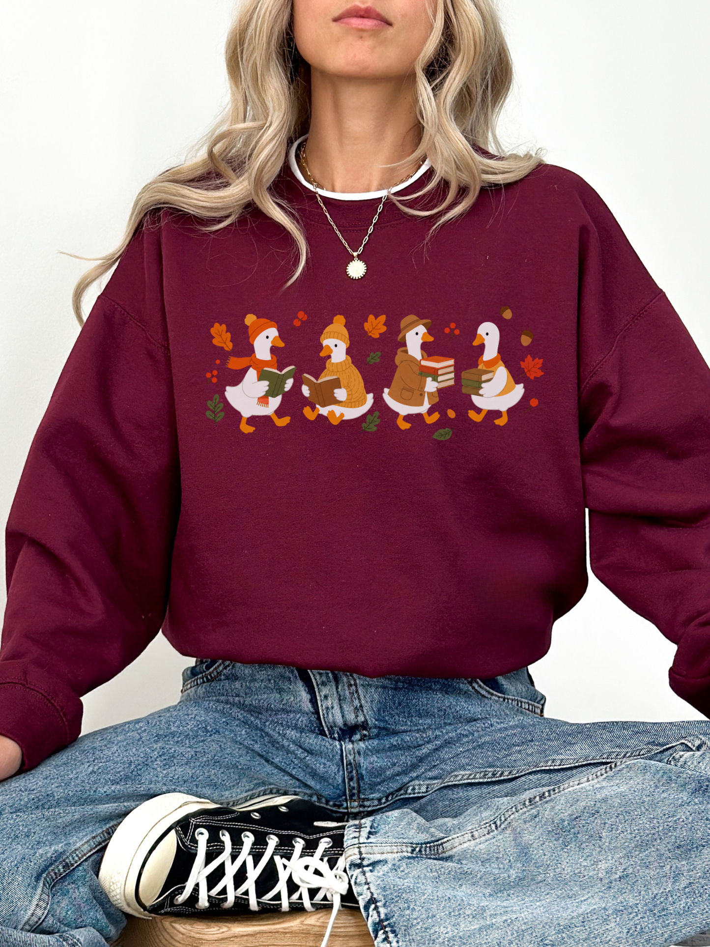 A woman wearing a maroon crewneck sweatshirt with a graphic of four cartoon ducks in autumn colors, paired with blue jeans and a black and white sneaker.
