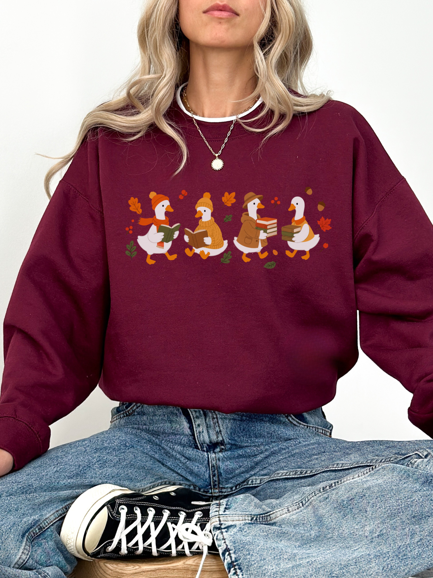 A woman wearing a maroon crewneck sweatshirt with a graphic of four cartoon ducks in autumn colors, paired with blue jeans and a black and white sneaker.