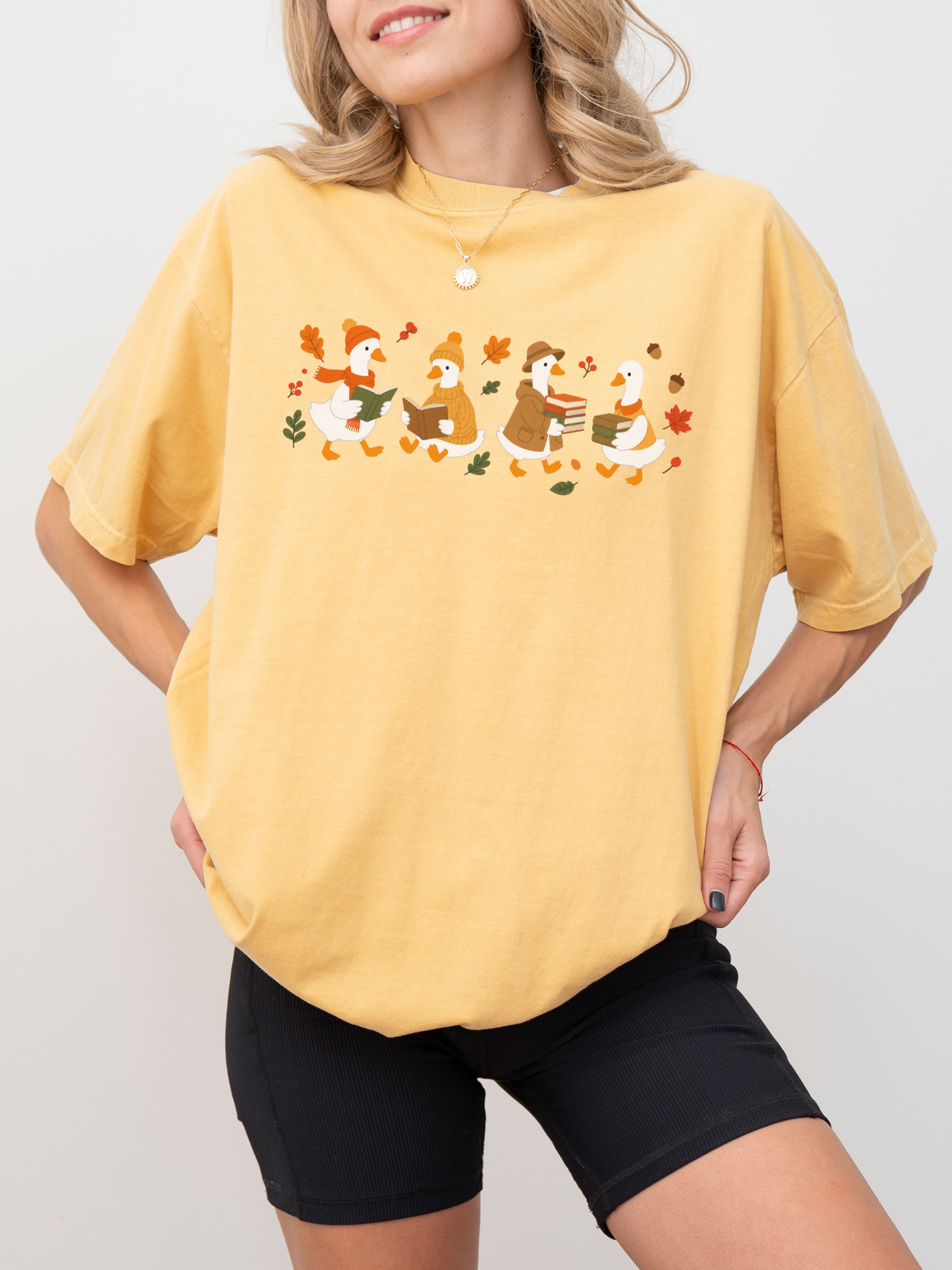 A woman wearing an oversized mustard t-shirt with a graphic of four cartoon ducks walking among autumn leaves, paired with black biker shorts against a light gray background.