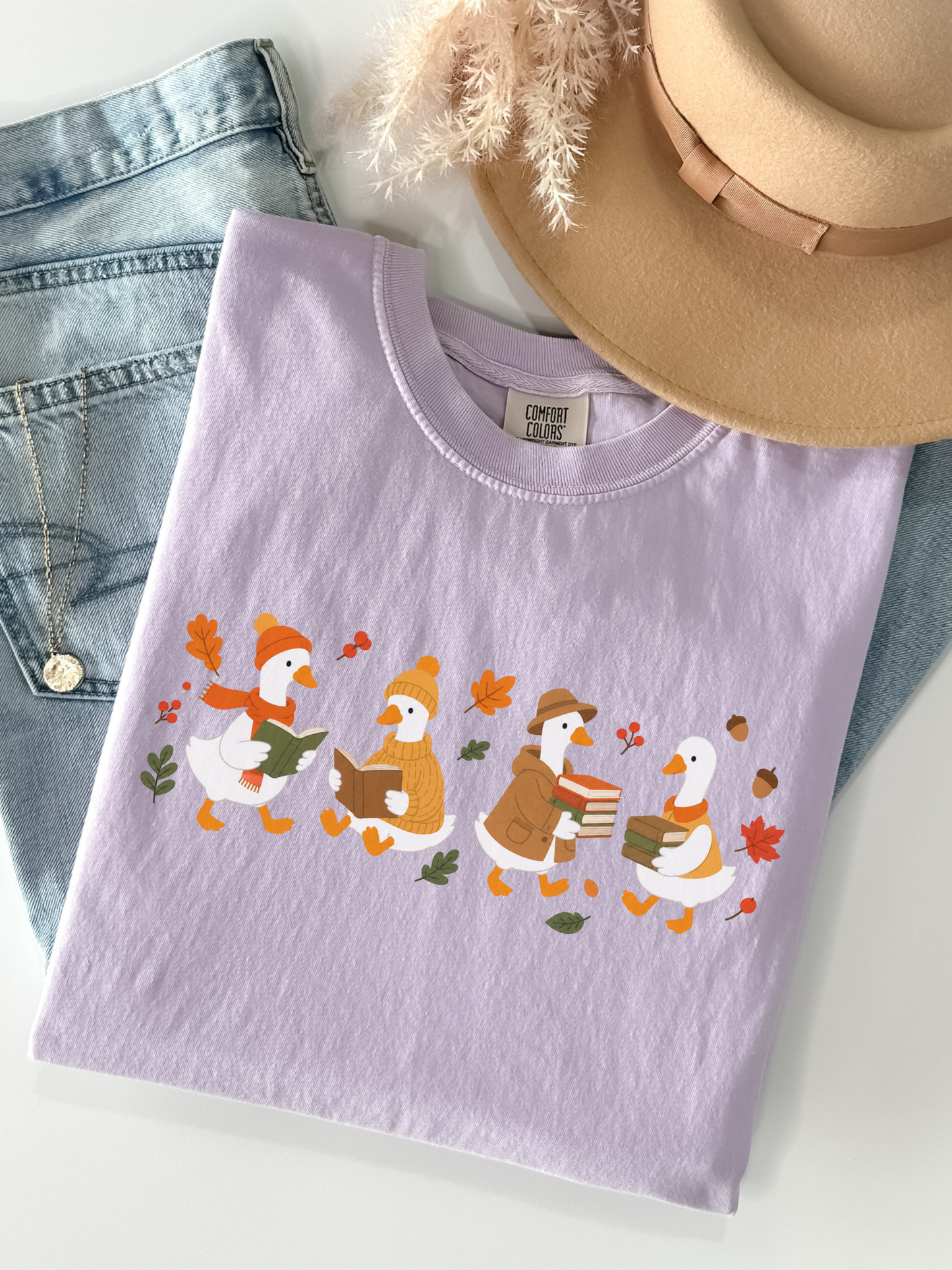 A folded light orchid crewneck t-shirt featuring a graphic of three reading ducks surrounded by autumn leaves, styled in a flat lay with blue jeans, a tan hat, and a necklace on a white background.