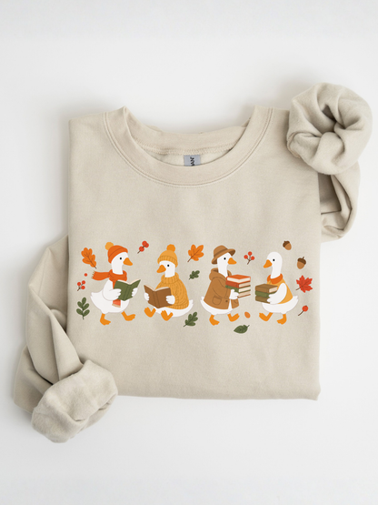 A folded beige crewneck sweatshirt featuring a graphic of four cartoon ducks reading books in autumn attire, styled with a matching beige scrunchie on a white background.