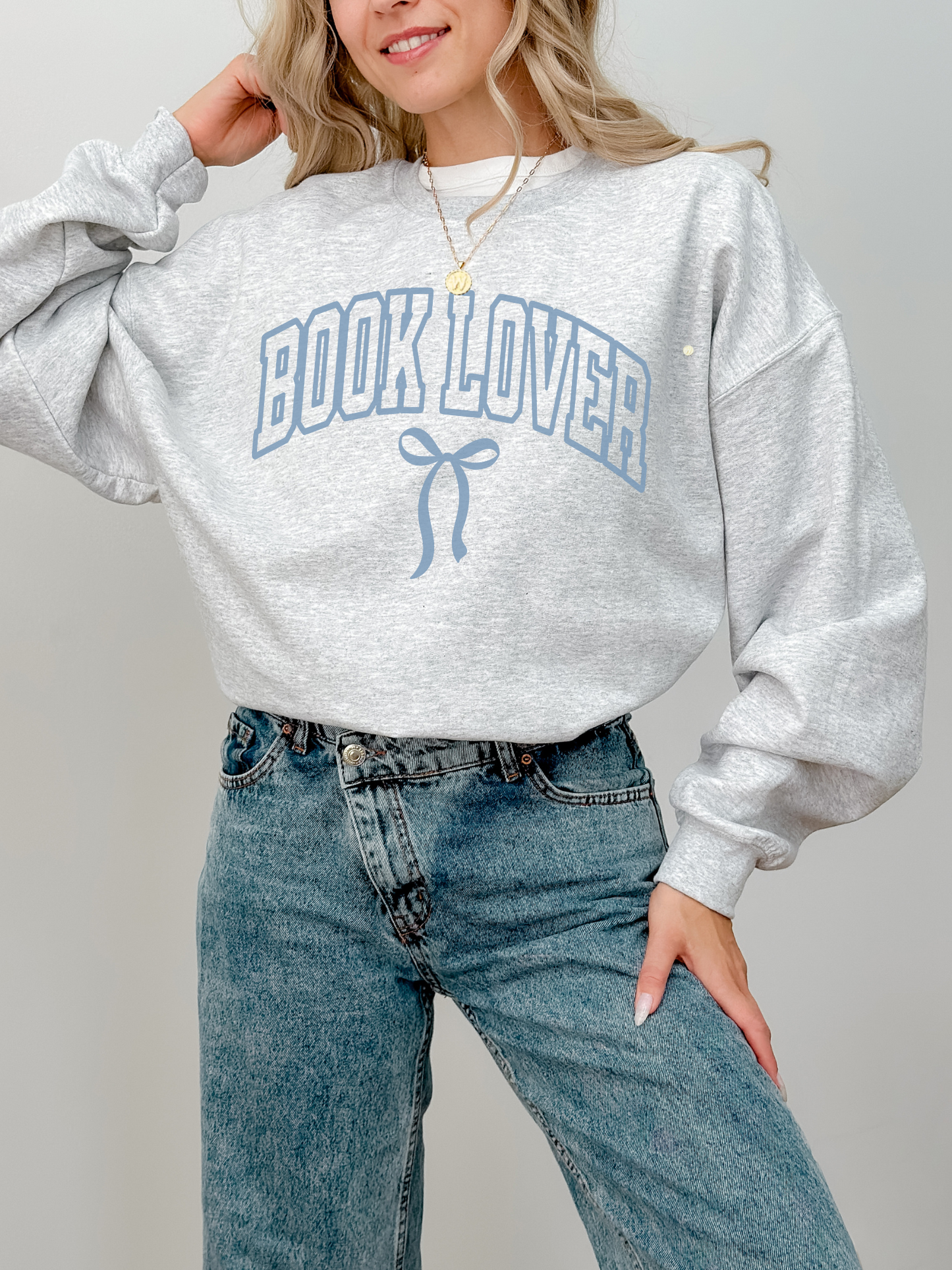 Woman wearing a light grey Book Lover varsity sweatshirt with a soft blue bow design, styled with jeans