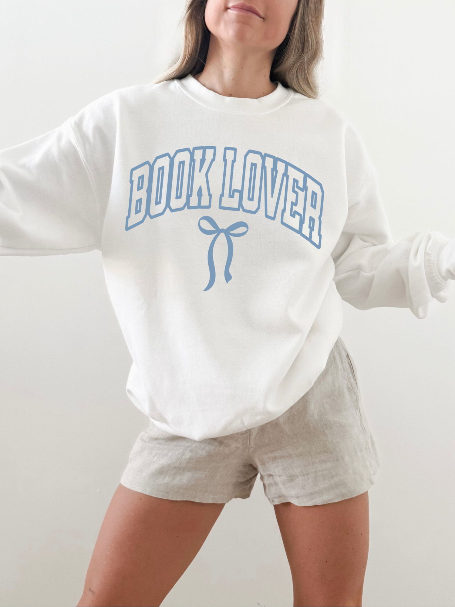 Woman in a white Book Lover varsity sweatshirt with blue bow, styled with beige shorts