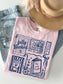 A light pink graphic T-shirt laid flat on a white surface, styled with blue jeans, a tan hat, and a necklace. The shirt has a navy blue book-themed print with phrases like 'fully Booked' and 'One more chapter.