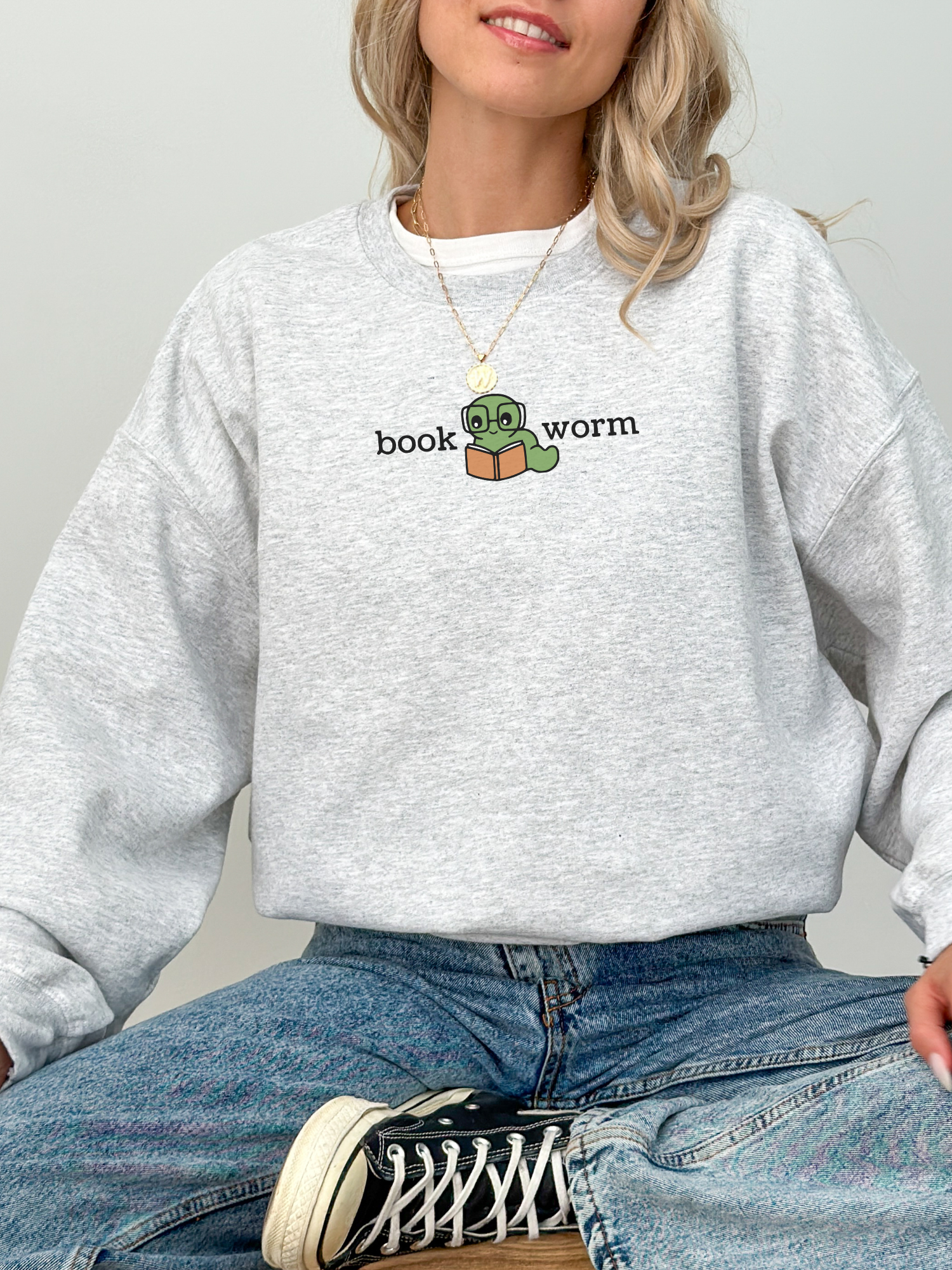 woman sitting cross-legged in an ash grey sweatshirt with a book worm design featuring a cute green worm and book