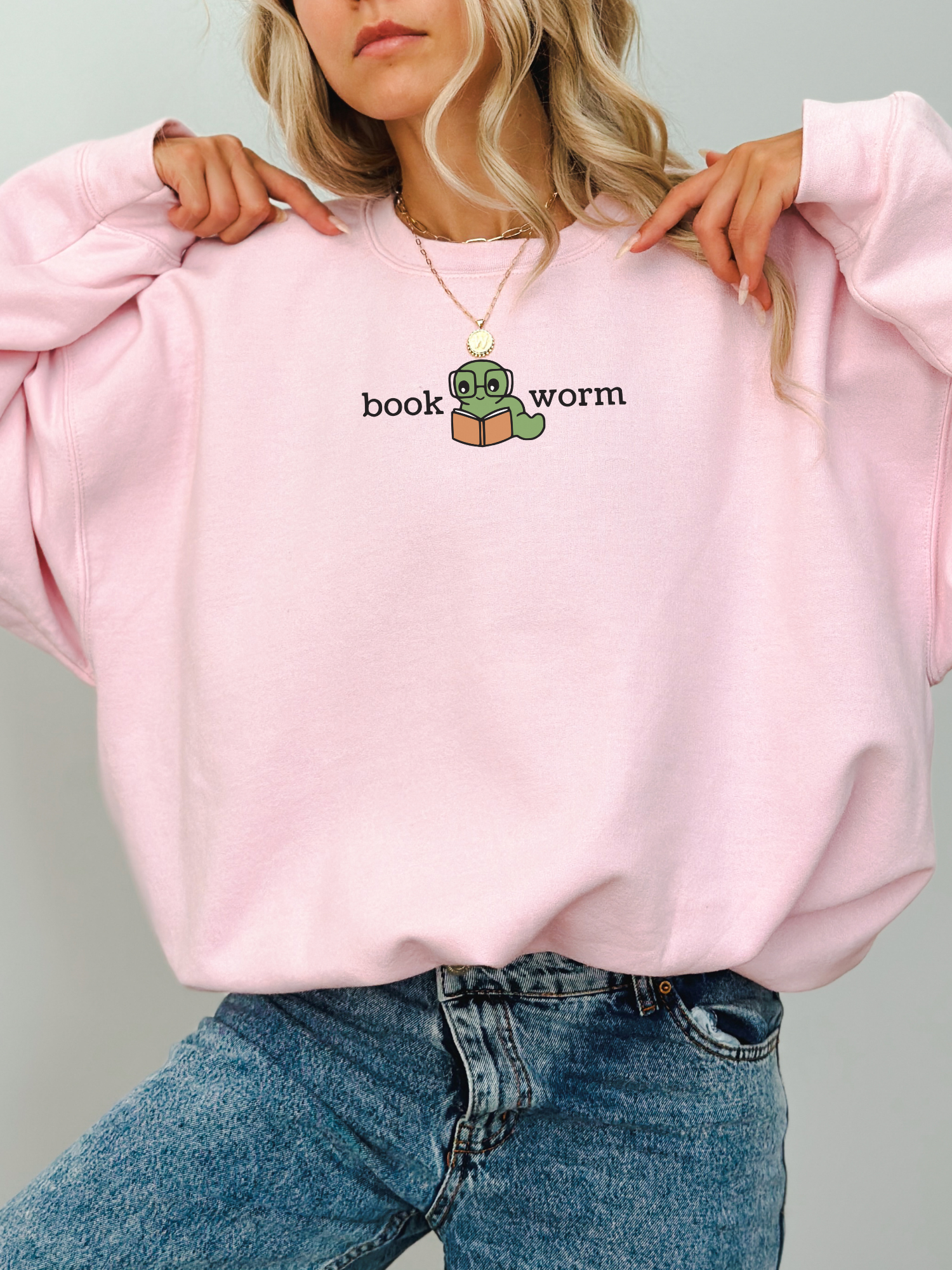 woman wearing a pink sweatshirt that says book worm with a cute green worm in glasses reading a book