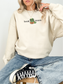 woman sitting in a sand-colored sweatshirt with book worm graphic and a cartoon worm reading an orange book