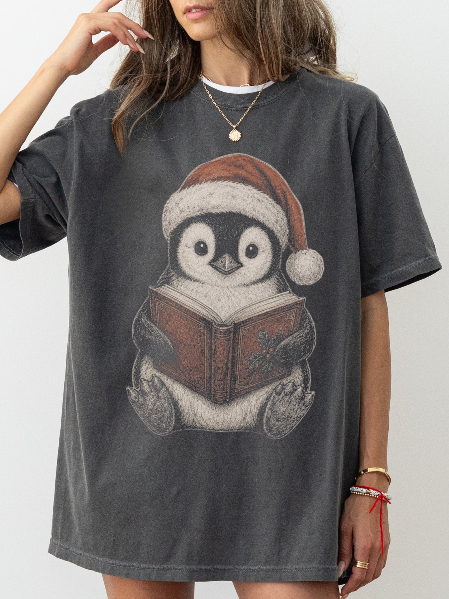 Model wearing a Pepper Comfort Colors bookish shirt with a Christmas penguin in a Santa hat reading a book with holly detail