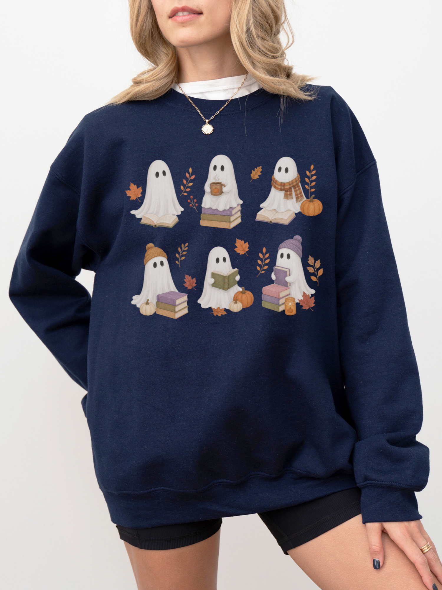 A woman wearing a navy blue crewneck sweatshirt with a graphic of six cute cartoon ghosts reading books in an autumn theme, paired with black bike shorts against a plain white background.