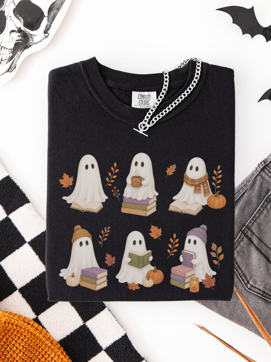 A folded black crewneck shirt with a graphic of six cute cartoon ghosts in autumn outfits, styled in a flat lay with a silver chain necklace, a skull, a checkered blanket, and denim.