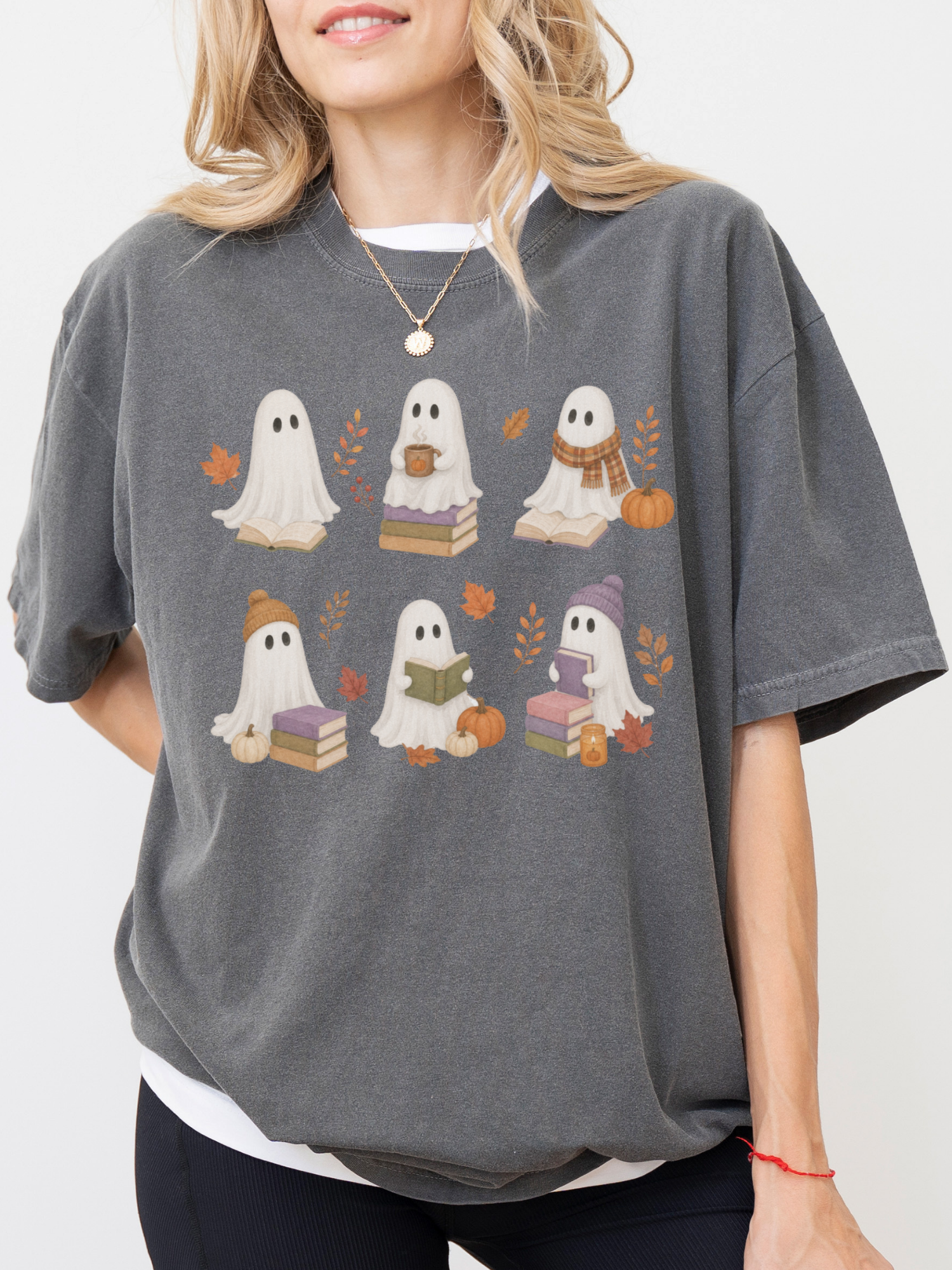 A woman wearing a pepper oversized t-shirt with a graphic print of six cute cartoon ghosts holding books and pumpkins, styled with a white undershirt.