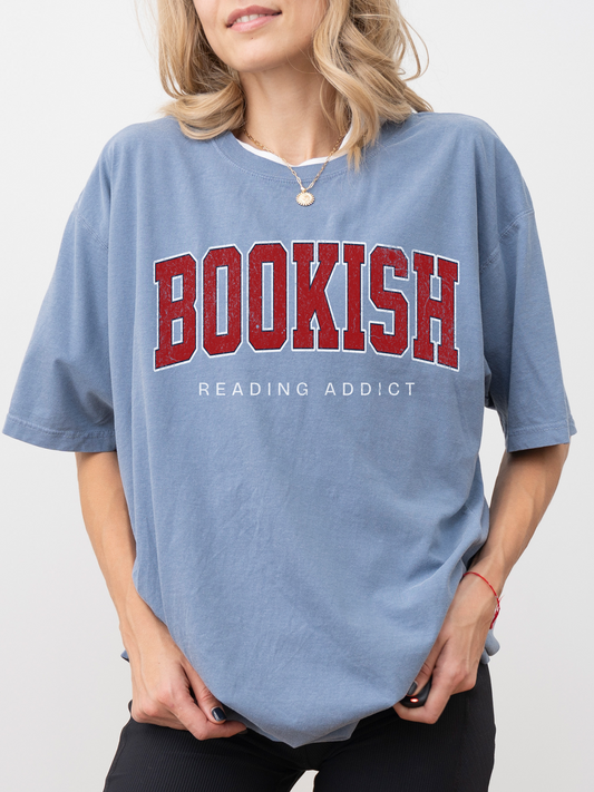 woman wearing blue Comfort Colors shirt that says Bookish Reading Addict in red varsity letters