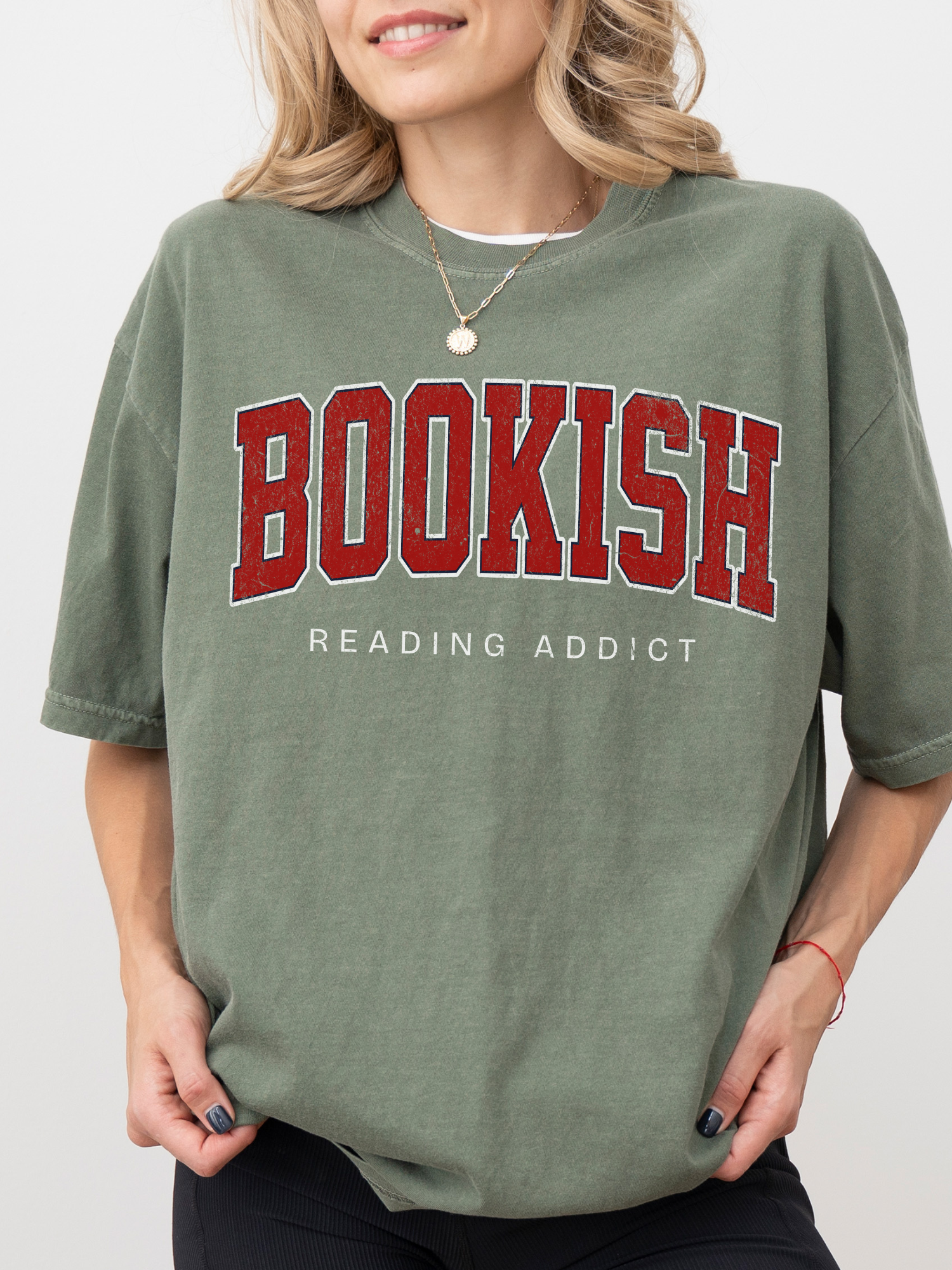woman wearing moss green Comfort Colors shirt that says Bookish Reading Addict in bold red varsity font