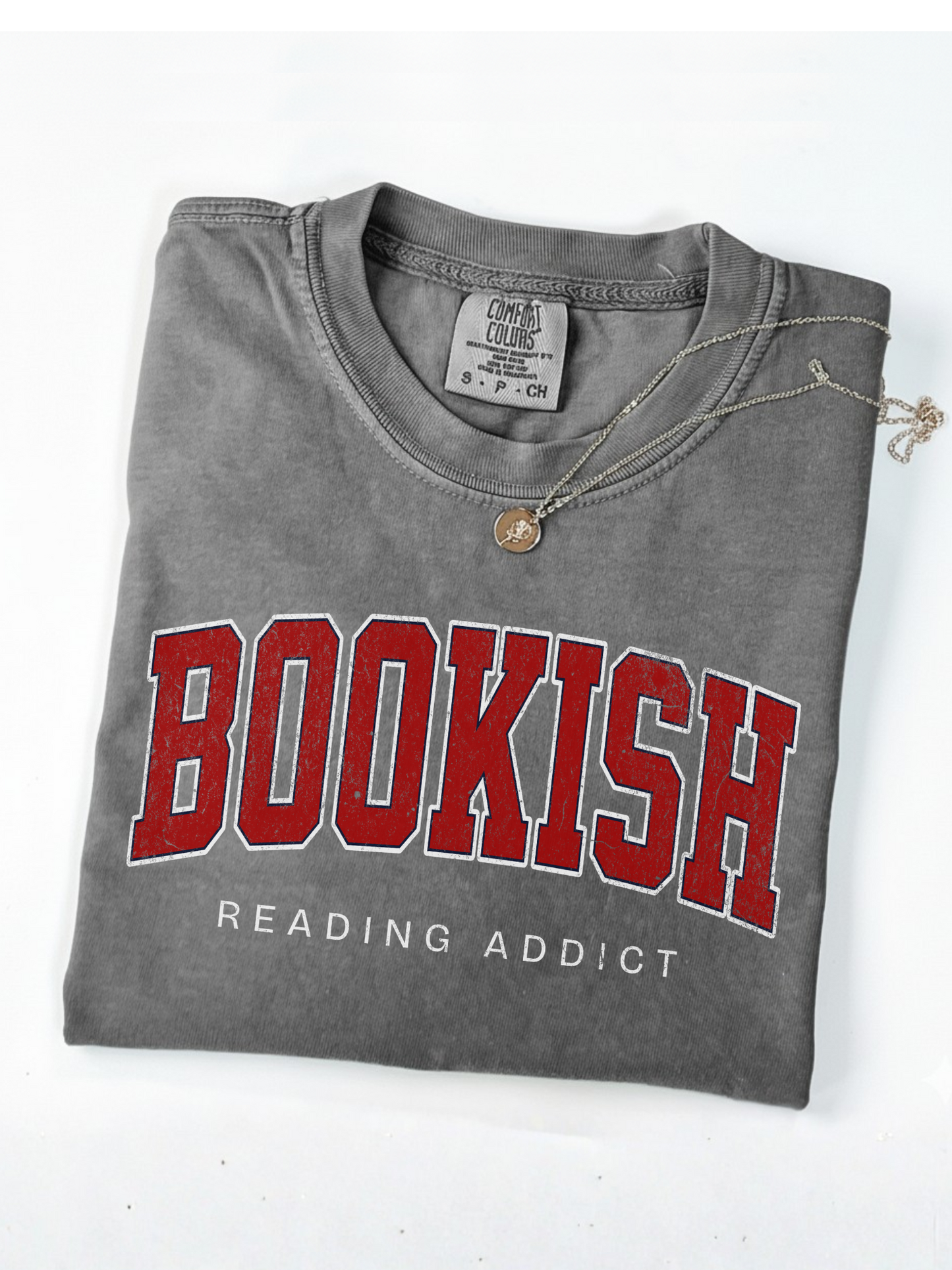 folded grey Comfort Colors shirt that says Bookish Reading Addict in red varsity letters with necklace