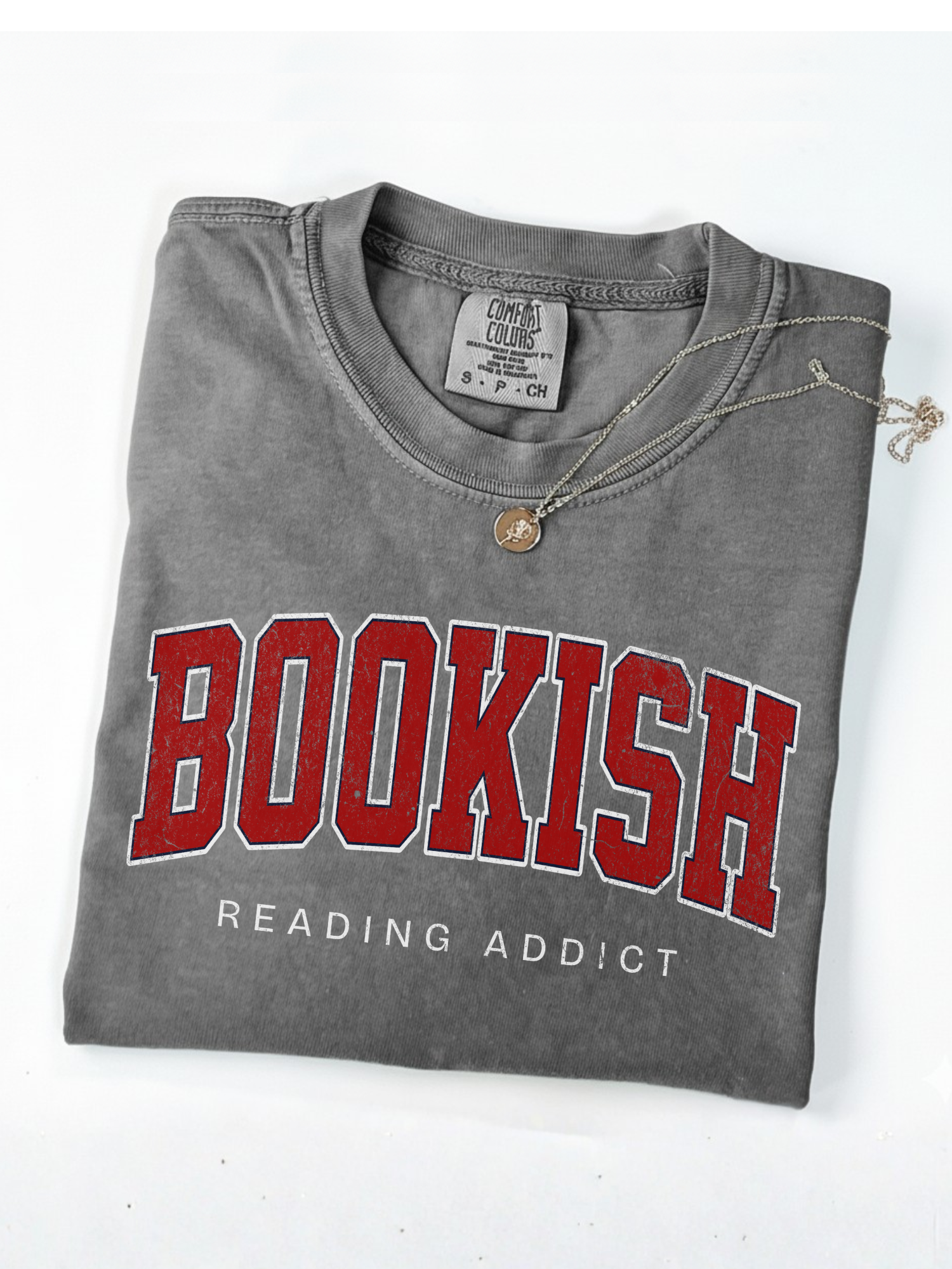 folded grey Comfort Colors shirt that says Bookish Reading Addict in red varsity letters with necklace