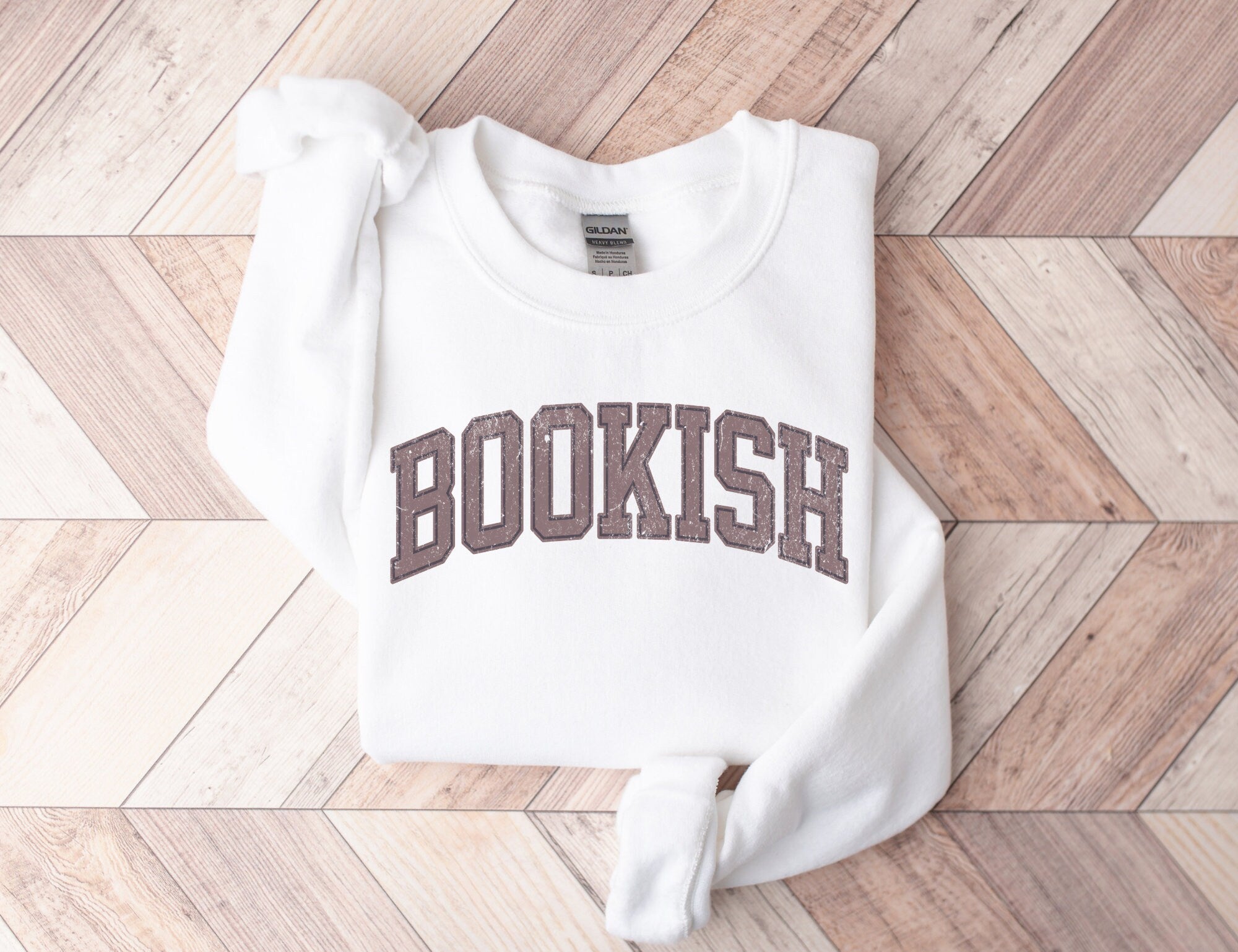 folded white sweatshirt with brown varsity-style bookish text on light wood herringbone background