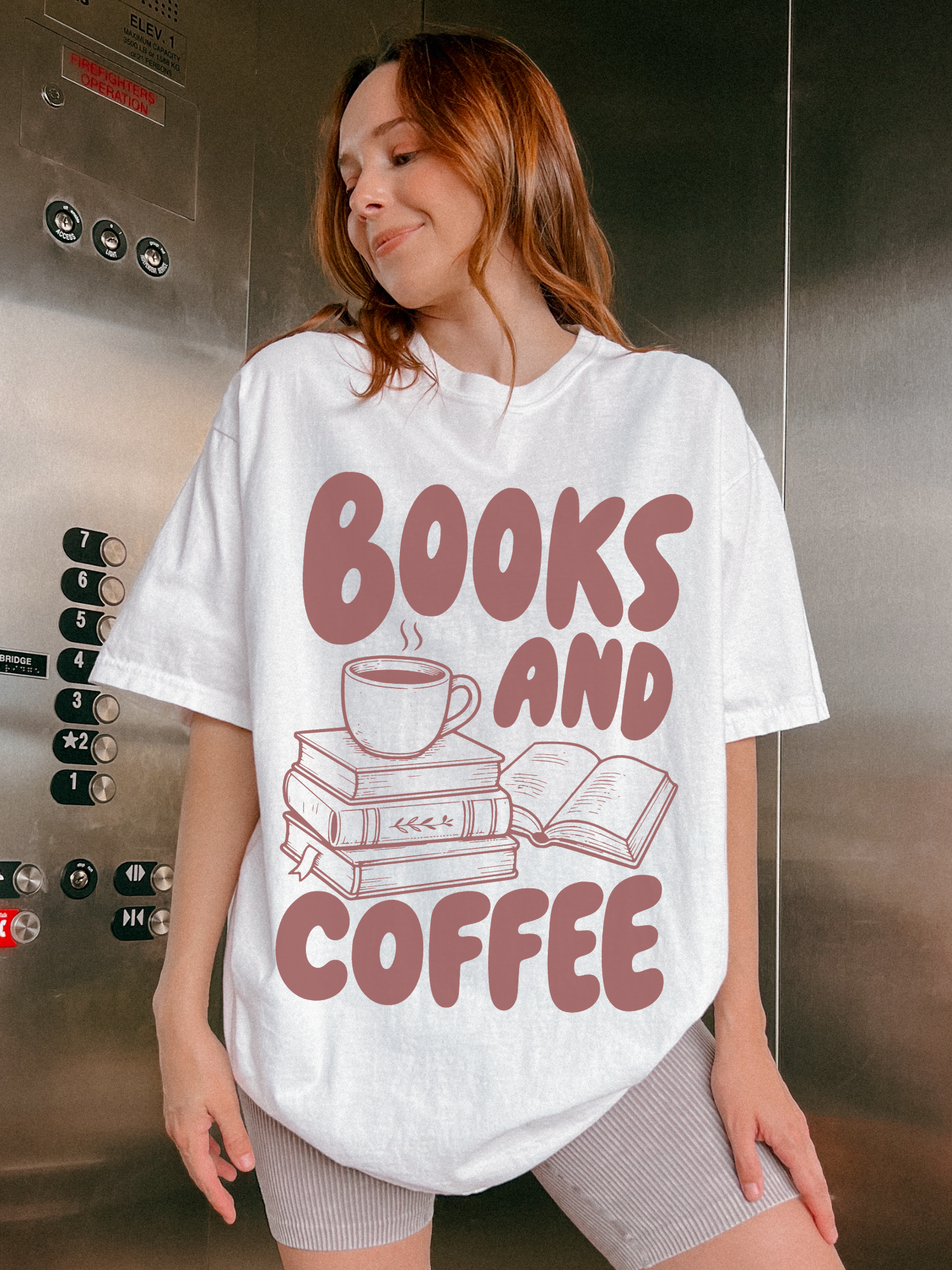 woman wearing a white Comfort Colors shirt featuring a books and coffee graphic with illustrated books and a steaming coffee cup, styled in an elevator setting for a cozy bookish aesthetic