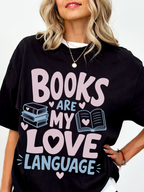 woman wearing a black comfort colors t shirt featuring books are my love language graphic with pastel pink and blue text stacked books open book illustration and heart details for book lovers