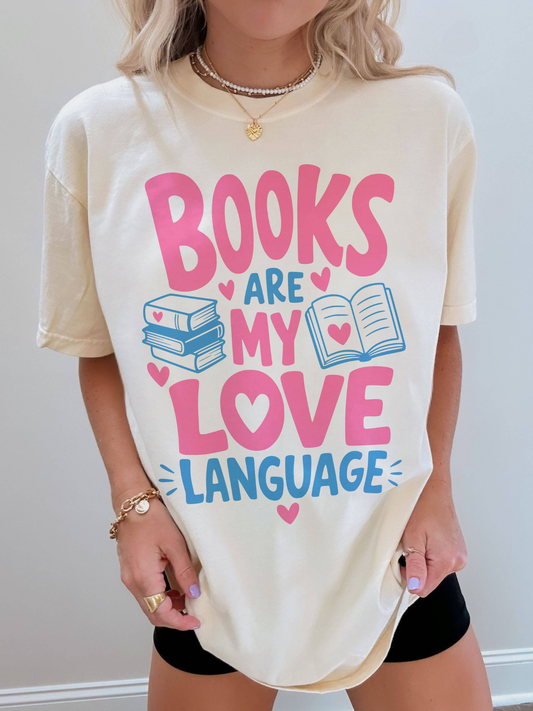woman wearing an ivory comfort colors t shirt featuring books are my love language design with pink and blue typography hearts stacked books and open book illustration for readers and book lovers