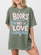 woman wearing a moss comfort colors t shirt featuring books are my love language graphic with stacked books open book illustration and pastel lettering for reading enthusiasts