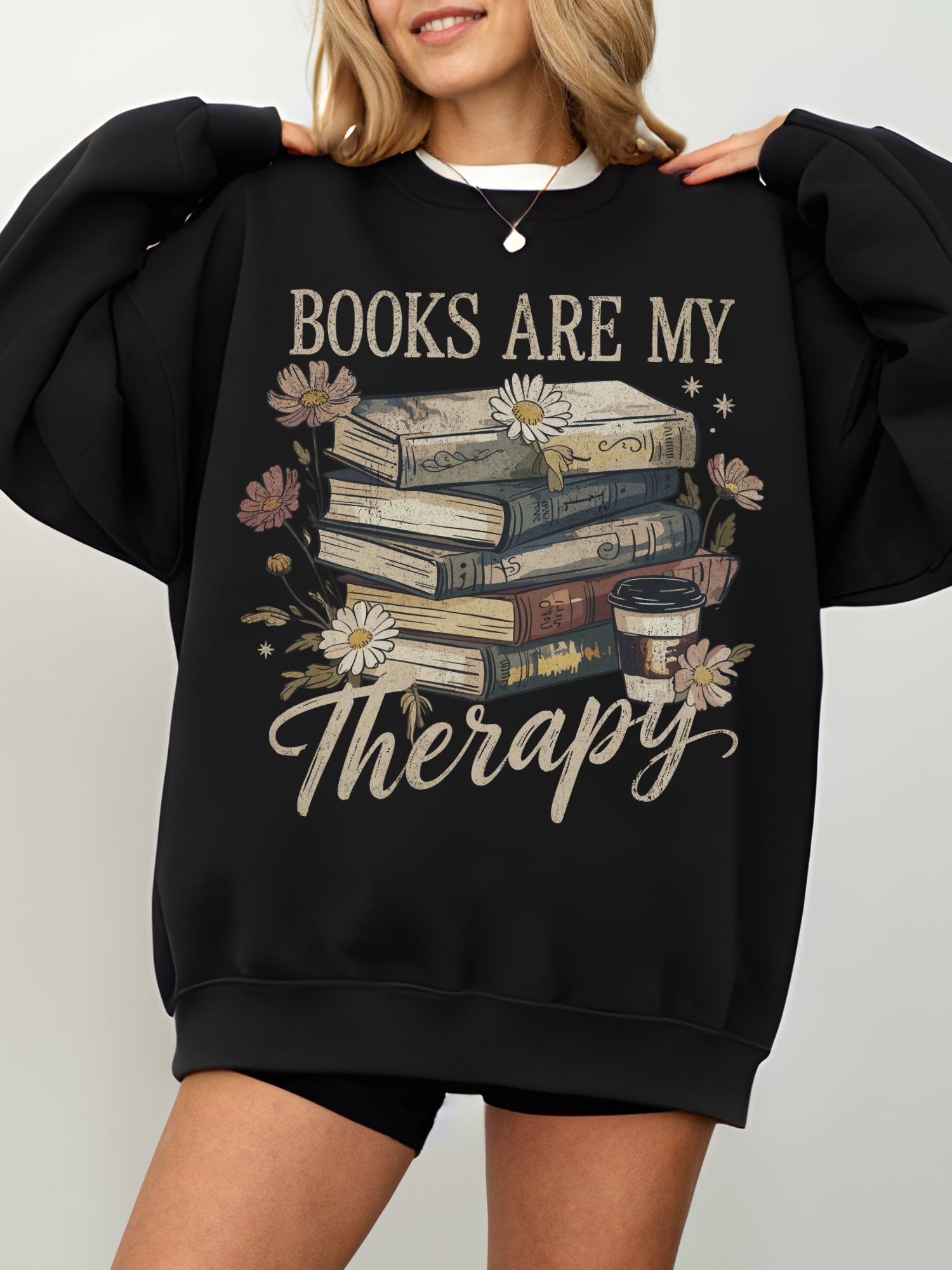 woman wearing oversized black sweatshirt with vintage floral books and coffee cup graphic that says books are my therapy