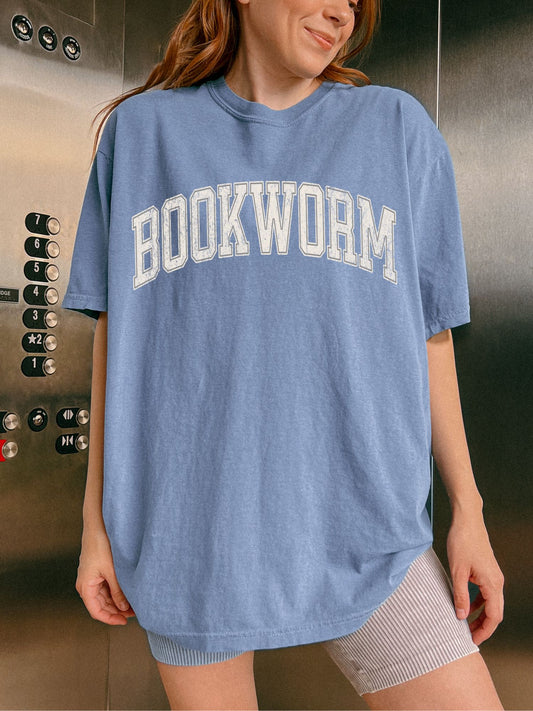 woman standing wearing a blue jean comfort colors shirt that says "bookworm" in a varsity college style font
