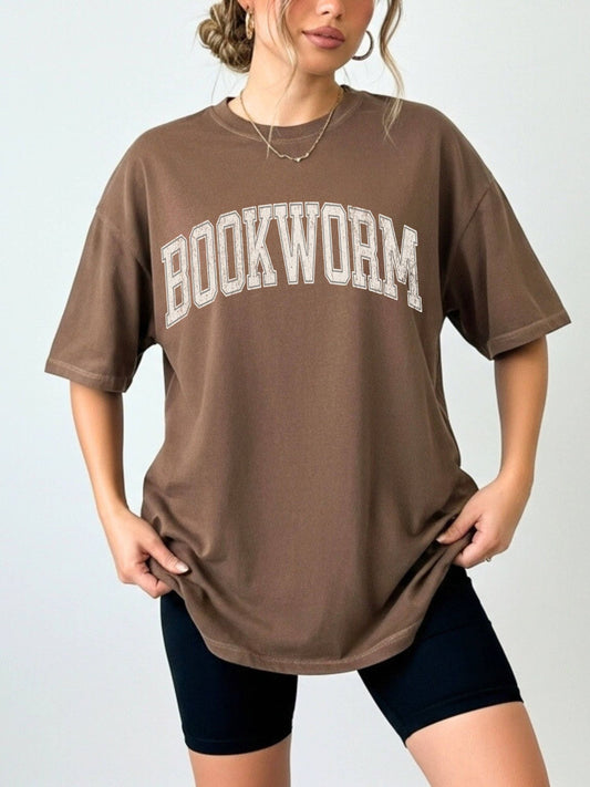woman standing wearing an espresso comfort colors shirt that says "bookworm" in a varsity college style font